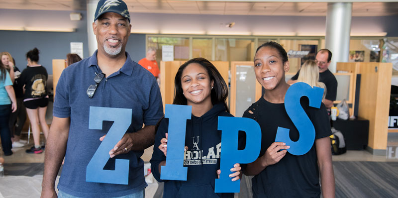 Parents of college student stand with her holding large blue letters that spell ZIPS