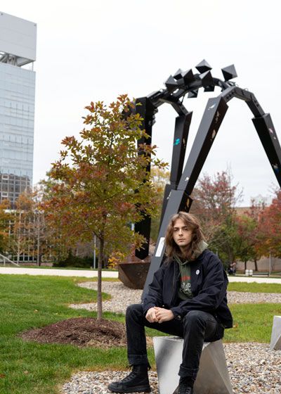 Young male student sitting outside in front of a 20-foot metal sculpture