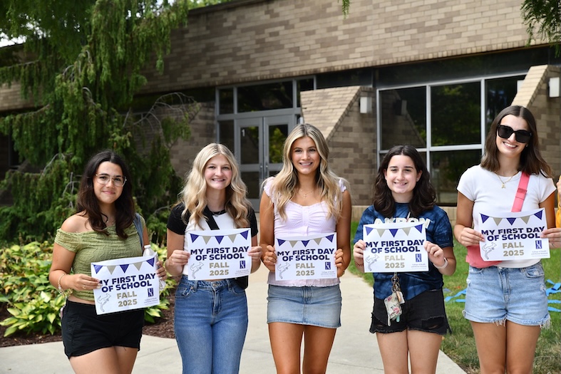 Five young women stand outside a building, smiling and holding signs that say My First Day of School Fall 2025. They are casually dressed and standing on a sidewalk with greenery in the background.