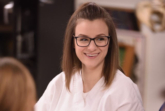 A young woman with straight brown hair and glasses smiles while wearing a white lab coat, sitting indoors with blurred objects in the background.