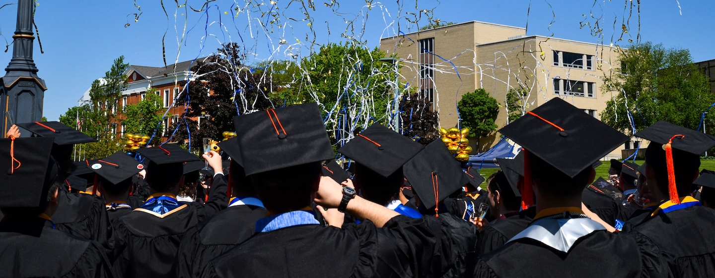 Graduates in caps and gowns celebrate outdoors under a blue sky, with silver streamers flying above them and a clock tower visible on the left. Buildings and green trees are in the background.