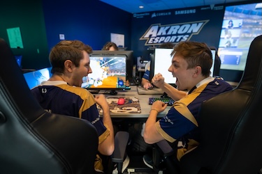 Two excited esports players in matching jerseys celebrate while playing a video game at computer desks in an Akron Esports gaming room.
