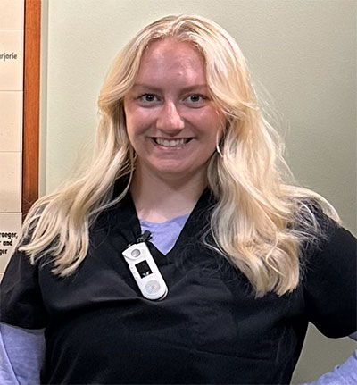 A woman with long blond hair wearing black medical scrubs and a badge smiles at the camera, standing indoors against a light-colored wall.