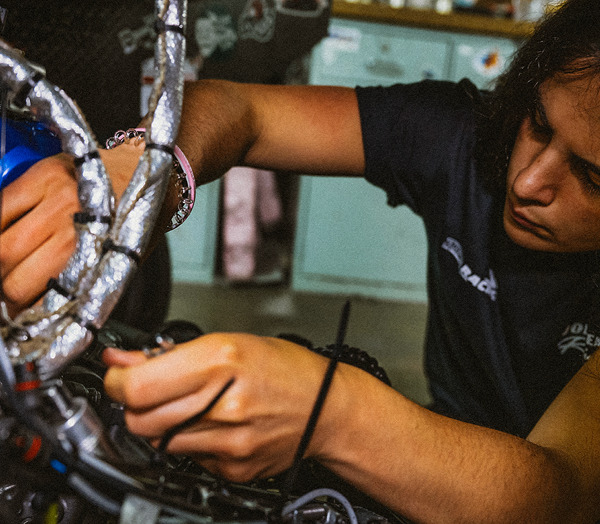 A person with long dark hair focuses intently as they work on wiring or assembling a mechanical device, holding tools and parts with both hands in a workshop setting.