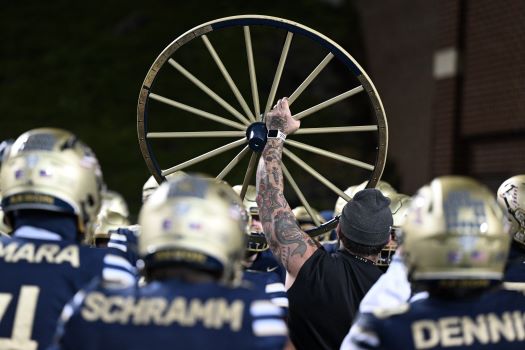 A tattooed arm raises a large wagon wheel above a group of football players in navy blue and gold uniforms, who stand close together with their backs to the camera.