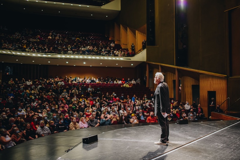 A person stands on a stage facing a large seated audience in a theater. The audience members appear engaged, and the theater is well-lit with several tiers of seating.