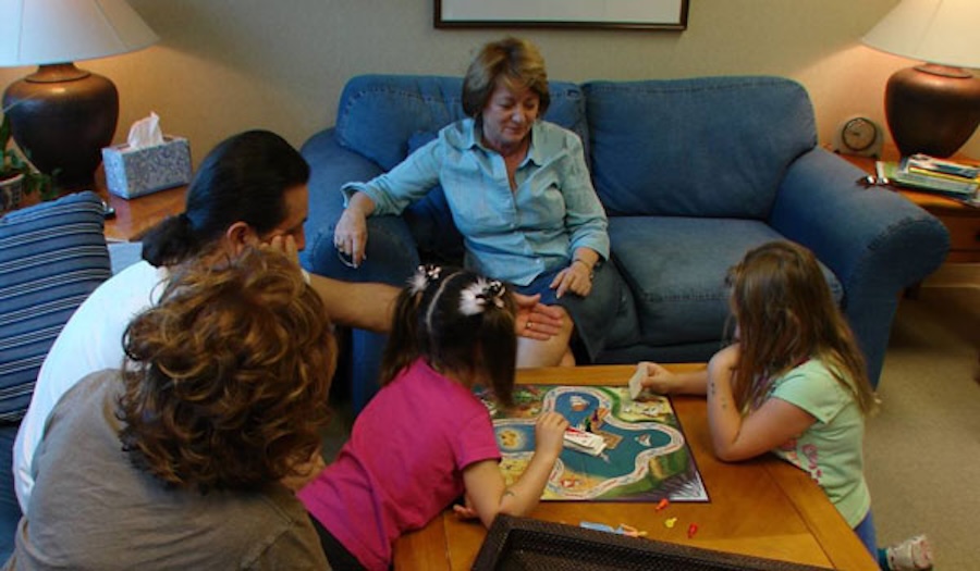 Four people sit around a wooden table playing a board game in a living room, while a woman sits on a blue couch behind them, watching and smiling.