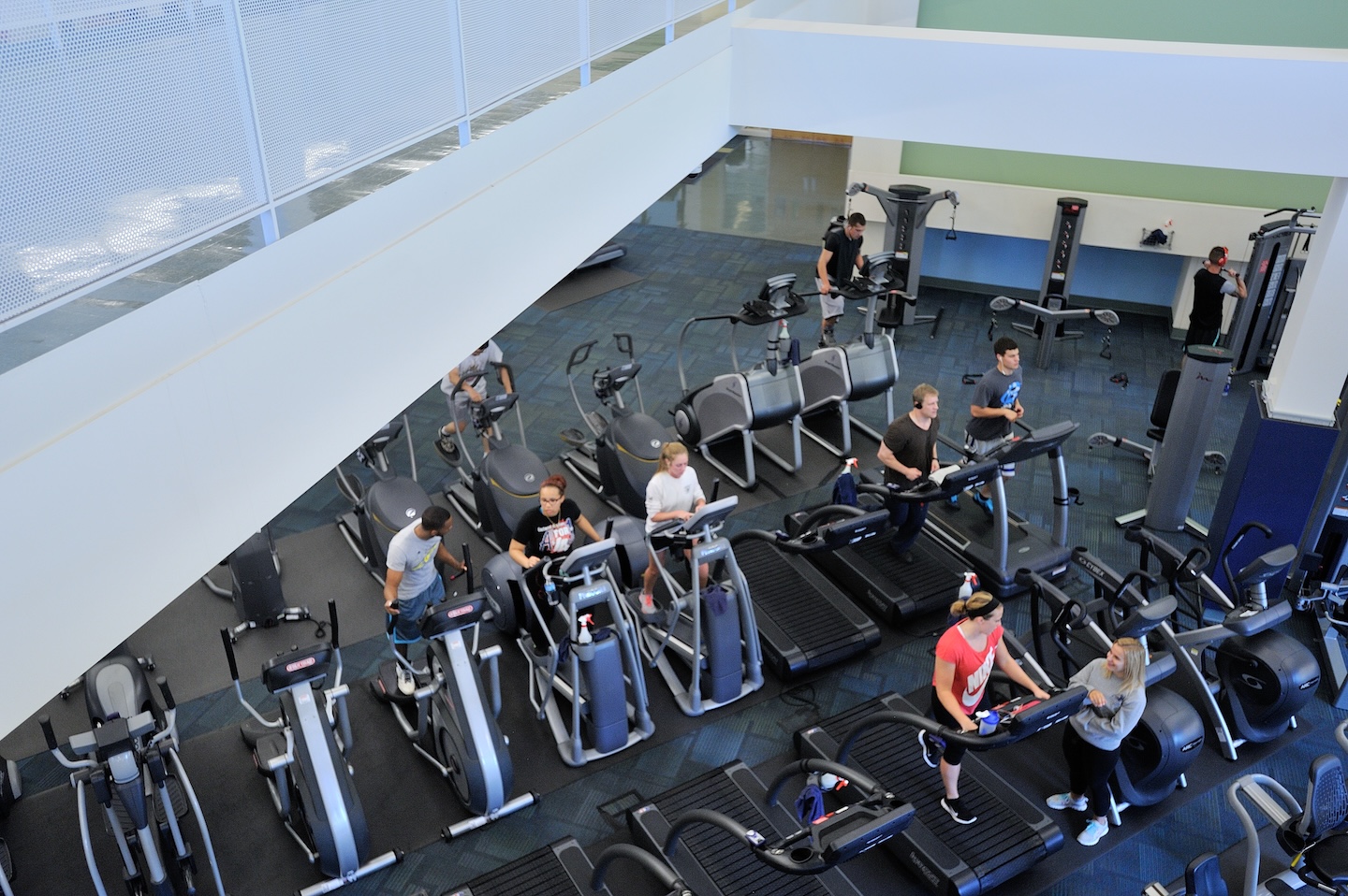 Students use treadmills, ellipticals, and weight machines in a modern, well-equipped campus recreation center fitness floor.