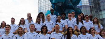 A group of young adults wearing matching white shirts pose together outdoors in front of a glass building with a large blue sculpture.