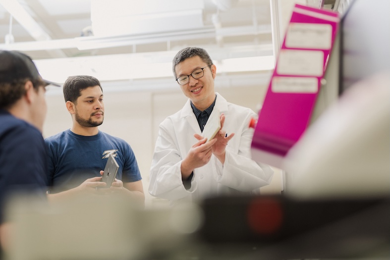 Three people stand in a laboratory. One, wearing a white lab coat and glasses, holds a rectangular object and explains something to the others, who are observing attentively. Equipment and machinery are visible in the foreground.