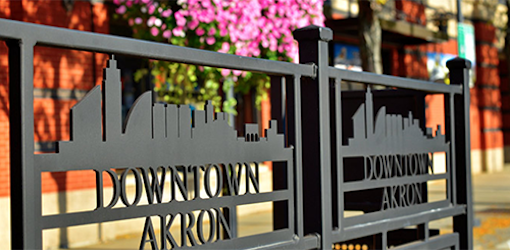 Black metal fence panels with a city skyline cutout and the words DOWNTOWN AKRON. Pink flowers and a brick building are visible in the background.