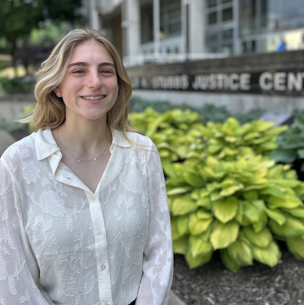 A young woman with long blond hair and a white blouse smiles while standing outside in front of green plants and a building with a sign that reads Justice Center.