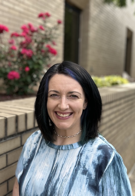 A woman with straight black hair and a blue-streaked top smiles while standing outside near a brick wall and blooming pink flowers.