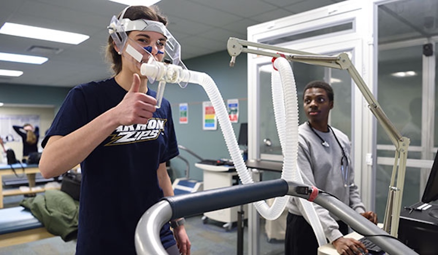 A person wearing a breathing mask connected to tubes walks on a treadmill, giving a thumbs up, while another person stands nearby monitoring equipment in a laboratory setting.