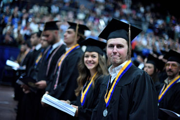 University of Akron graduates in caps and gowns stand in a row during a commencement ceremony inside a large arena.