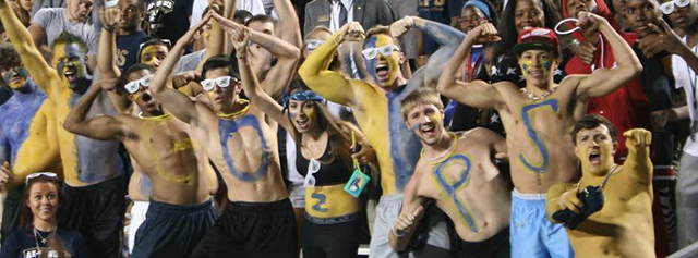 A group of enthusiastic sports fans, many with faces and bodies painted in blue and yellow, cheer and pose shirtless in the stands. Some have letters painted on their chests, and a festive atmosphere surrounds them.