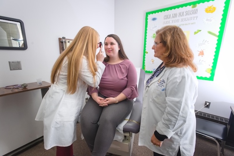 A woman sits on an exam table while a healthcare professional examines her eyes with an ophthalmoscope as another professional observes.