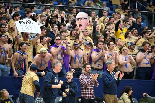 A large group of enthusiastic basketball fans in yellow and blue cheer in stadium stands; some have painted chests spelling AM I BLUE ZIPS, and hold a sign reading FEAR ZIPPY and a large cutout of a bald mans face.