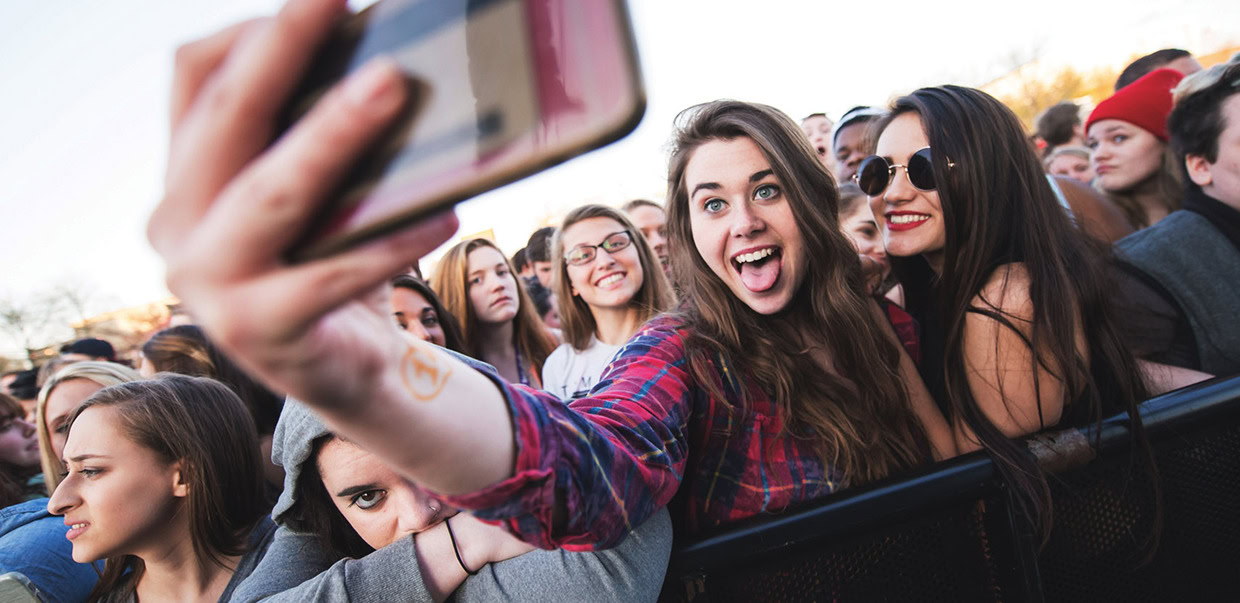 A group of young people stands at an outdoor event, with one person in the foreground taking a selfie and others smiling behind her.