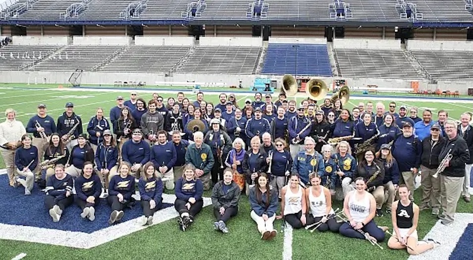 A large marching band group poses on a football field, with band members holding instruments and wearing uniforms. The stadium seats in the background are mostly empty.