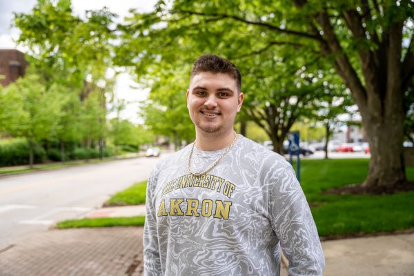 A young man stands outdoors on a sidewalk, smiling at the camera. He wears a gray sweatshirt with The University of Akron printed in yellow letters. Green trees and a street are visible in the background.