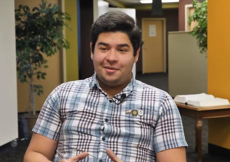 A young man with dark hair wearing a plaid shirt sits indoors, speaking with his hands. There is a plant and a table with an open book behind him, and office doors are visible in the background.