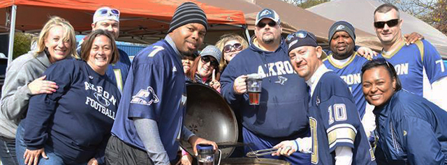 A group of people wearing Akron football jerseys gather outdoors, smiling and posing together beside a grill at a tailgate event under tents.