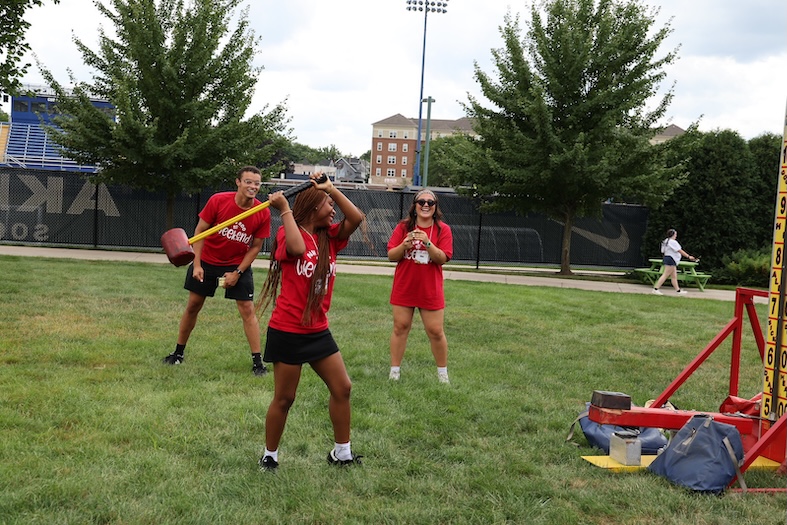 Three people wearing red shirts play a high striker game outdoors. One person swings a mallet, while two others stand behind her, smiling and watching. Trees, grass, and sports field fencing are visible in the background.