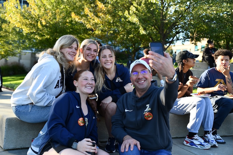A group of five young women and one adult man pose for a selfie outdoors on what appears to be a college campus. The man, wearing a white cap and glasses, holds up a smartphone to take the photo. The women, dressed in navy and gray sweatshirts featuring an 'A' logo, lean in and smile. Trees with autumn foliage are visible in the background, along with other students seated nearby.