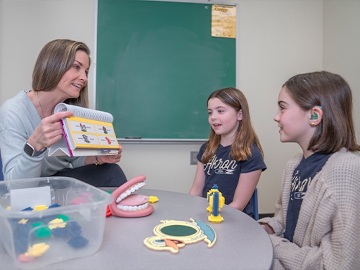 A woman shows a book with visual aids to two young girls, one of whom wears a hearing aid. Educational tools like a model ear and mouth are on the table in a classroom setting with a green chalkboard in the background.