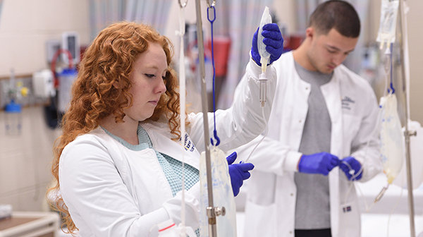 Two healthcare professionals in white coats and purple gloves prepare IV bags in a clinical setting, with medical equipment visible in the background.