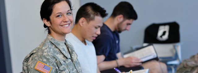 A woman in military uniform smiles at the camera while two men next to her focus on writing and reading. They are seated indoors, and the setting appears to be a classroom or study space.