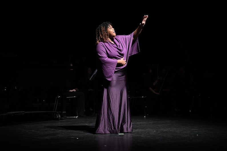 A performer in a long, purple dress stands on a dark stage, one arm raised dramatically, as if reaching out, while the other rests across their body. A spotlight highlights the performer, with the background mostly in shadow.