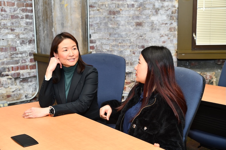 Two women sit together at a wooden conference table in conversation. The woman on the left, wearing a black blazer over a green turtleneck, smiles and rests her chin on her hand while listening. The woman on the right, wearing a black jacket, faces her and speaks. Both are seated in blue office chairs against a backdrop of exposed brick walls.