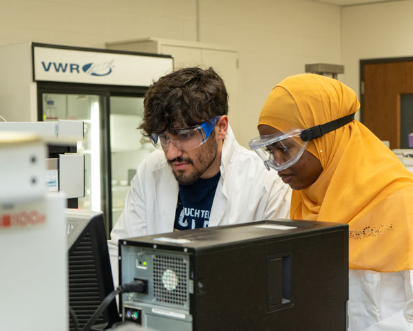 Two scientists wearing lab coats and safety goggles work together at a computer in a laboratory setting.