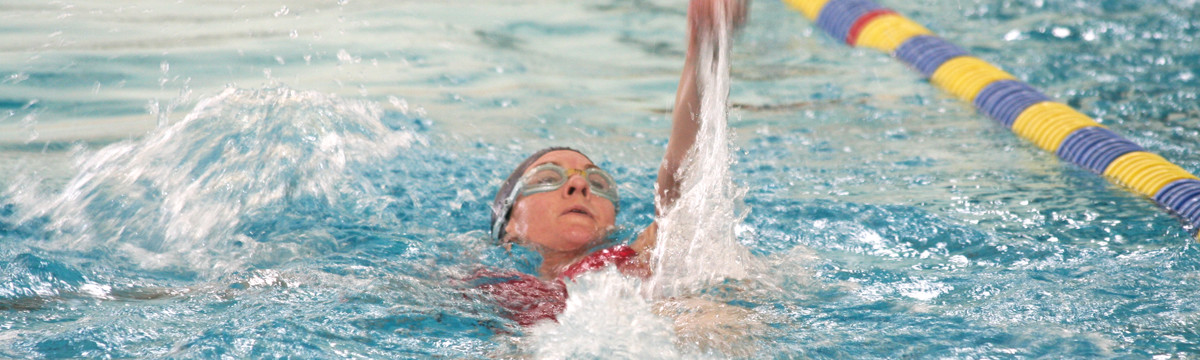 A swimmer wearing goggles does the backstroke in an indoor campus pool.
