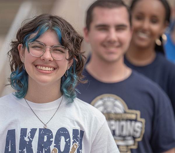 University of Akron student with blue-streaked hair smiles in the foreground while classmates stand behind on campus.