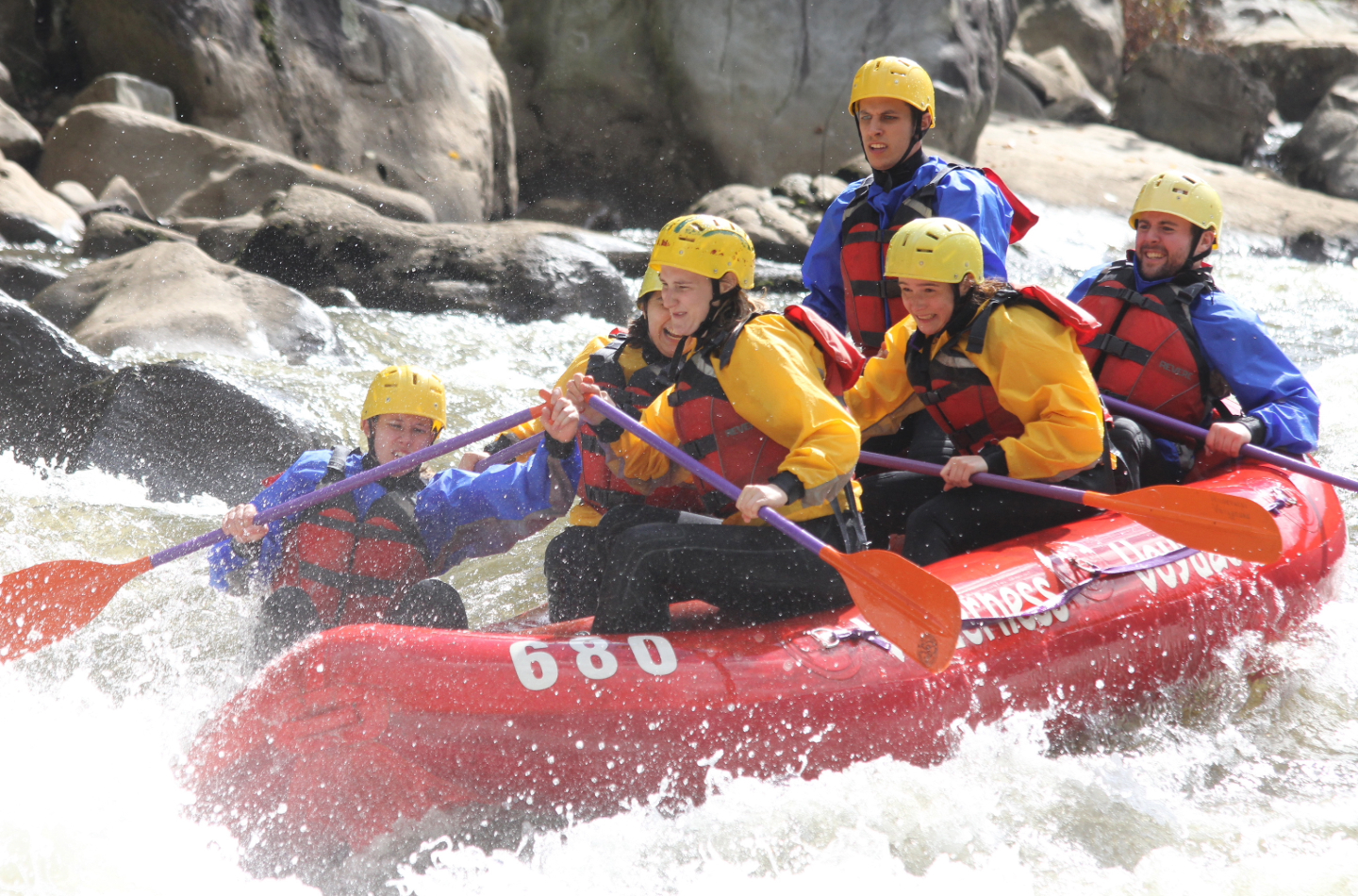 Five people in helmets and life jackets paddle a red inflatable raft through whitewater rapids, splashing water around them as they navigate the river surrounded by rocks.