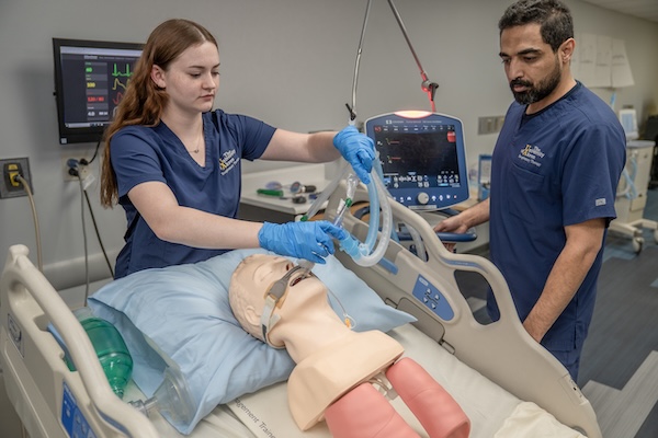 Two healthcare professionals in blue scrubs practice placing a respiratory mask on a medical mannequin in a hospital bed, with medical monitors and equipment visible in the background.