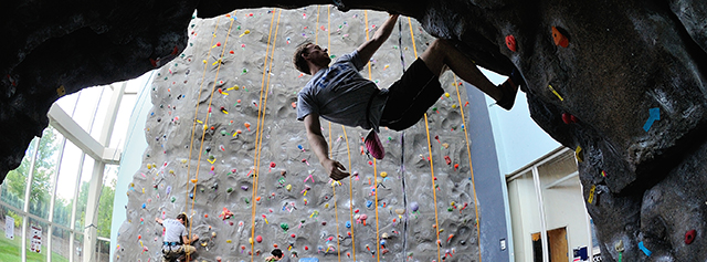 A person climbs on an indoor rock wall, hanging from an overhang, while others climb in the background on vertical climbing routes with colorful holds.