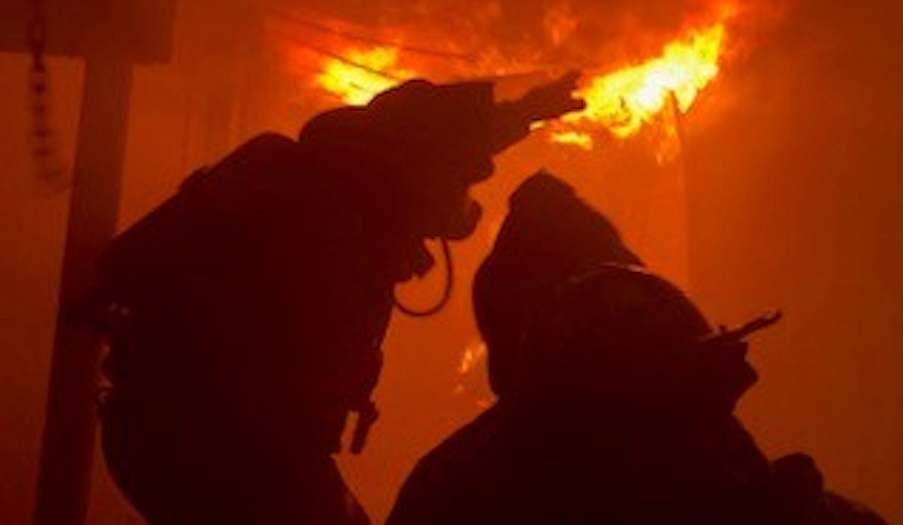 Two firefighters in protective gear are silhouetted against intense orange flames inside a burning building, reaching upward as they work to control the fire.