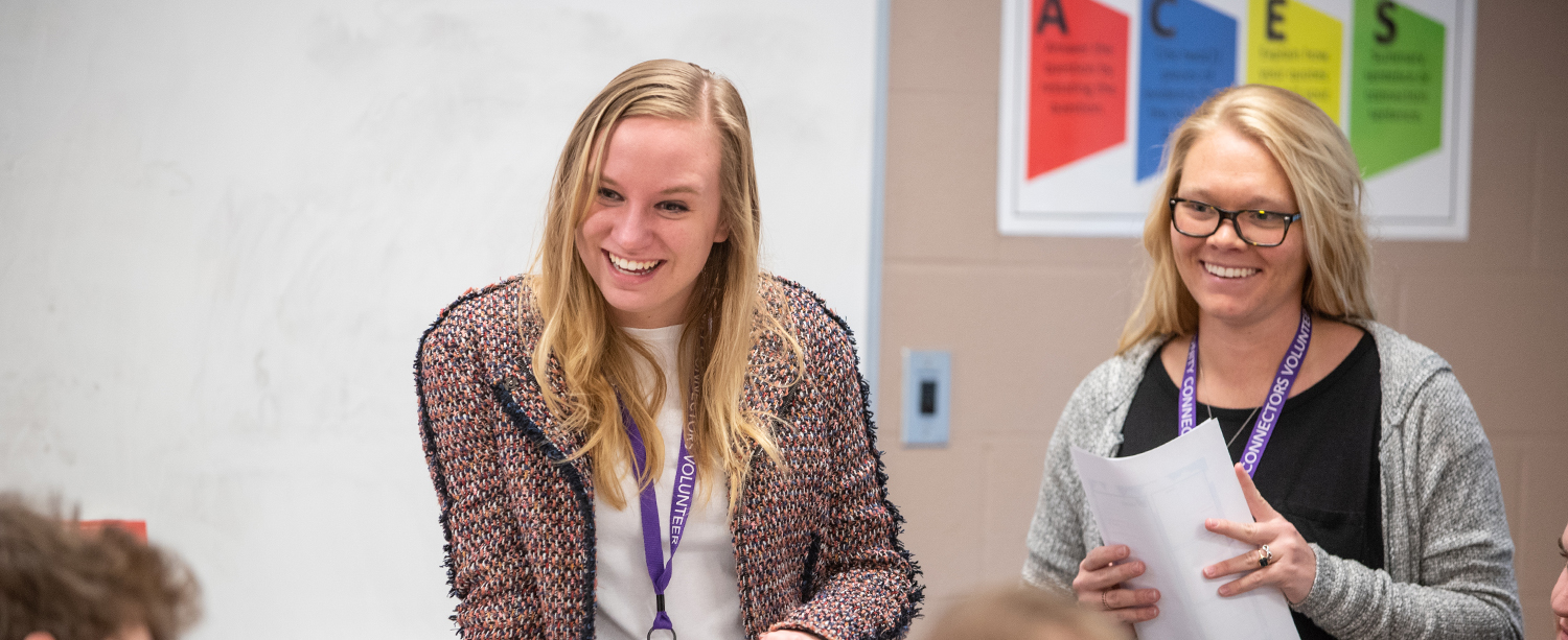 Two women wearing lanyards smile and stand in front of a classroom whiteboard, one holding papers. A colorful educational poster is visible on the wall behind them.