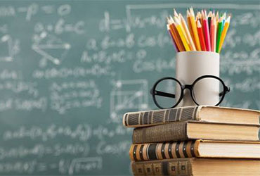 Stack of books, pencils and eyeglasses in front of a classroom chalkboard, representing education and teaching careers.