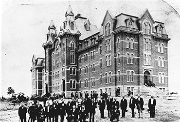 Black and white photo of an early 20th-century campus street lined with parked vintage cars and surrounded by large brick buildings with towers and chimneys in the background.