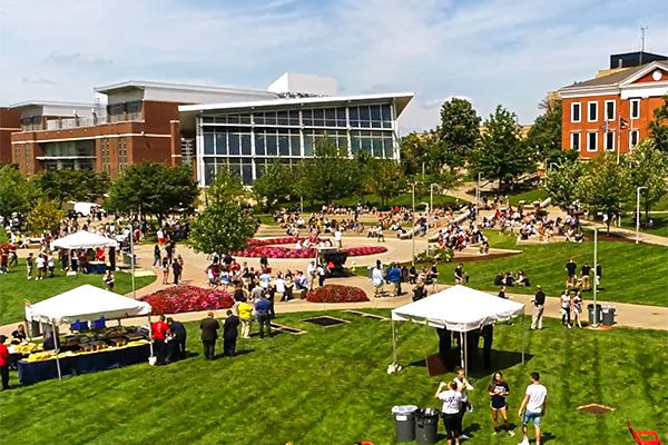 A high-angle view of a bustling college campus common area on a sunny day. Numerous people are gathered on a large green lawn and surrounding concrete pathways, with several white event tents set up. Modern and traditional brick university buildings sit in the background under a blue sky with light clouds.