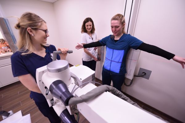 Two women in scrubs prepare another woman for a chest X-ray; one aligns the X-ray machine while the patient stands with arms outstretched, and another woman smiles and observes.