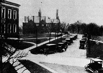 Black and white photo of a campus walkway between two buildings, with a person walking in the center and another in the background. Beyond the walkway, there are fields and a few distant trees.