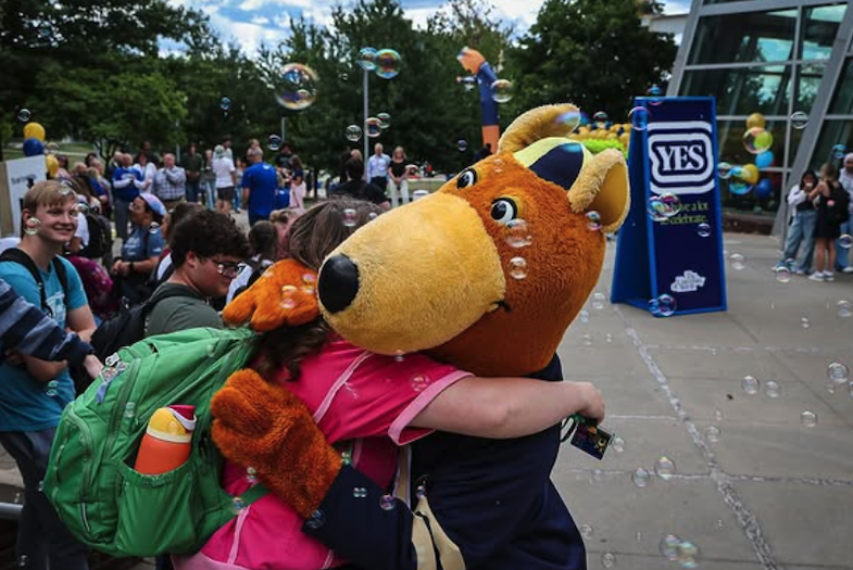 A person in a kangaroo mascot costume hugs a smiling student outdoors as bubbles float around. Other people watch and smile near a building with trees in the background.