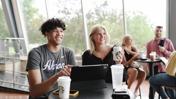 Two smiling students sit together at a table inside a brightly lit campus building with large glass windows. The male student, wearing a grey 