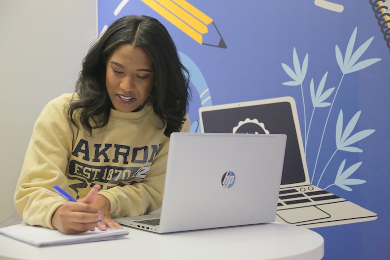 A woman in an Akron University sweatshirt writes in a notebook with a pen while sitting at a table with an open HP laptop. A colorful illustrated background with school supplies and plants is behind her.